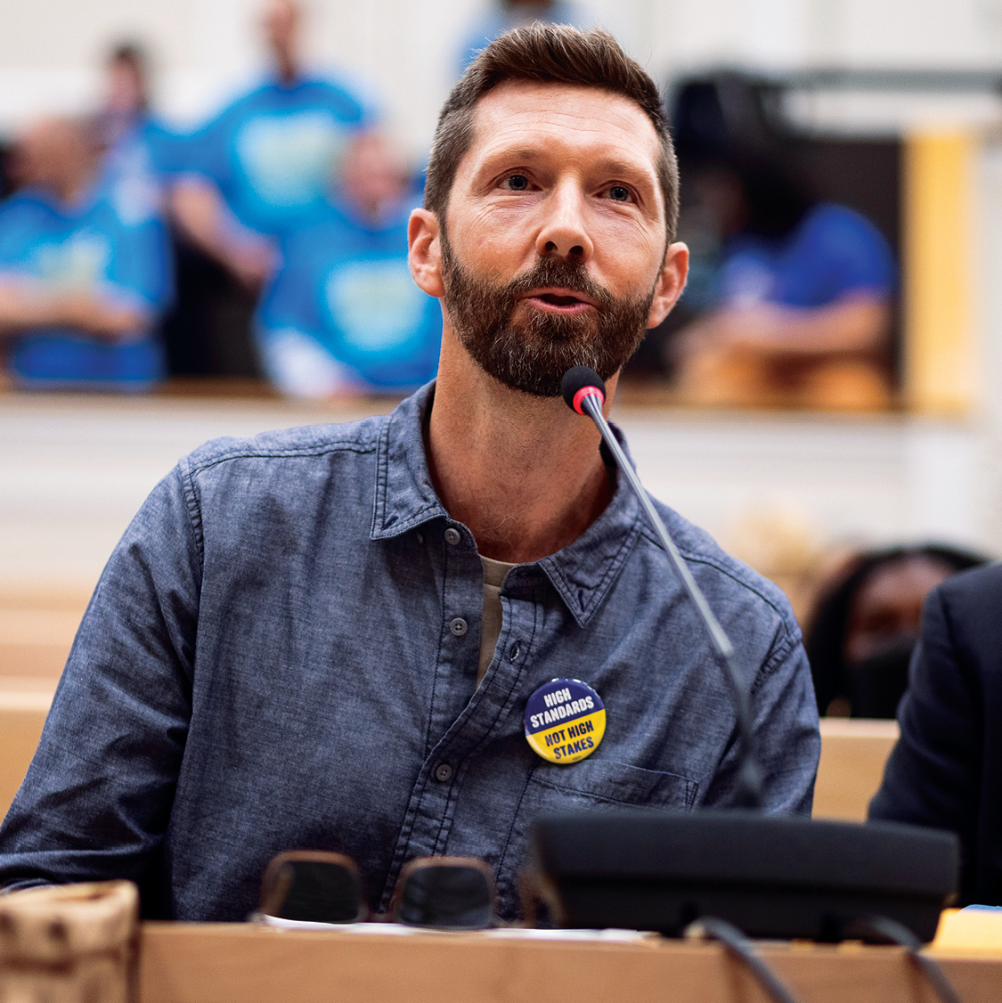 A white man with a beard in a blue shirt speaking at a microphone in a meeting