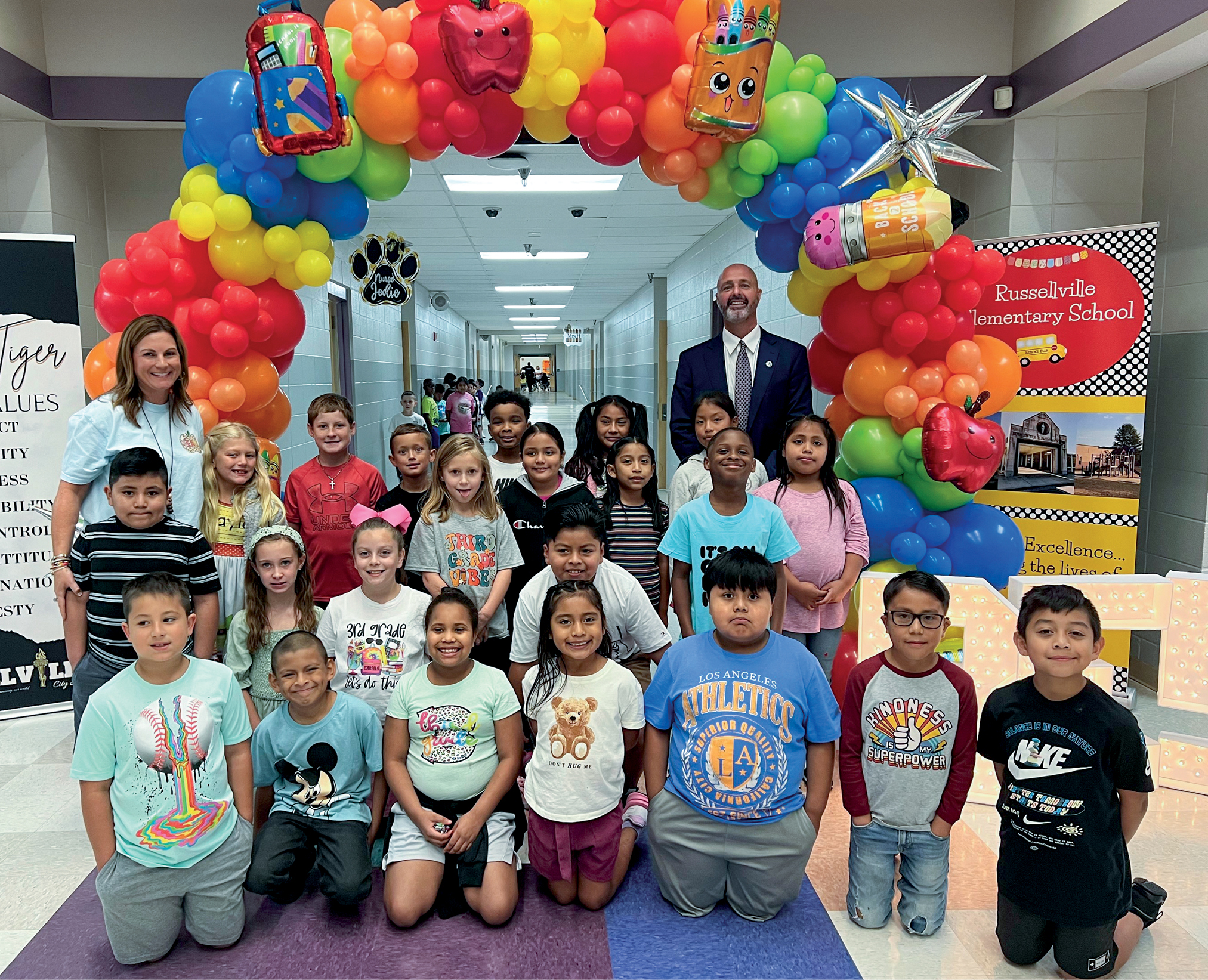 A group of students sitting and posing underneath a balloon arch with two adults
