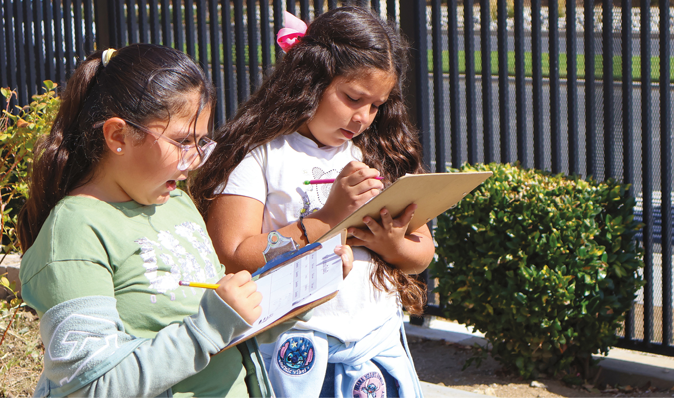 Two elementary aged girls taking notes on clipboards in their school garden