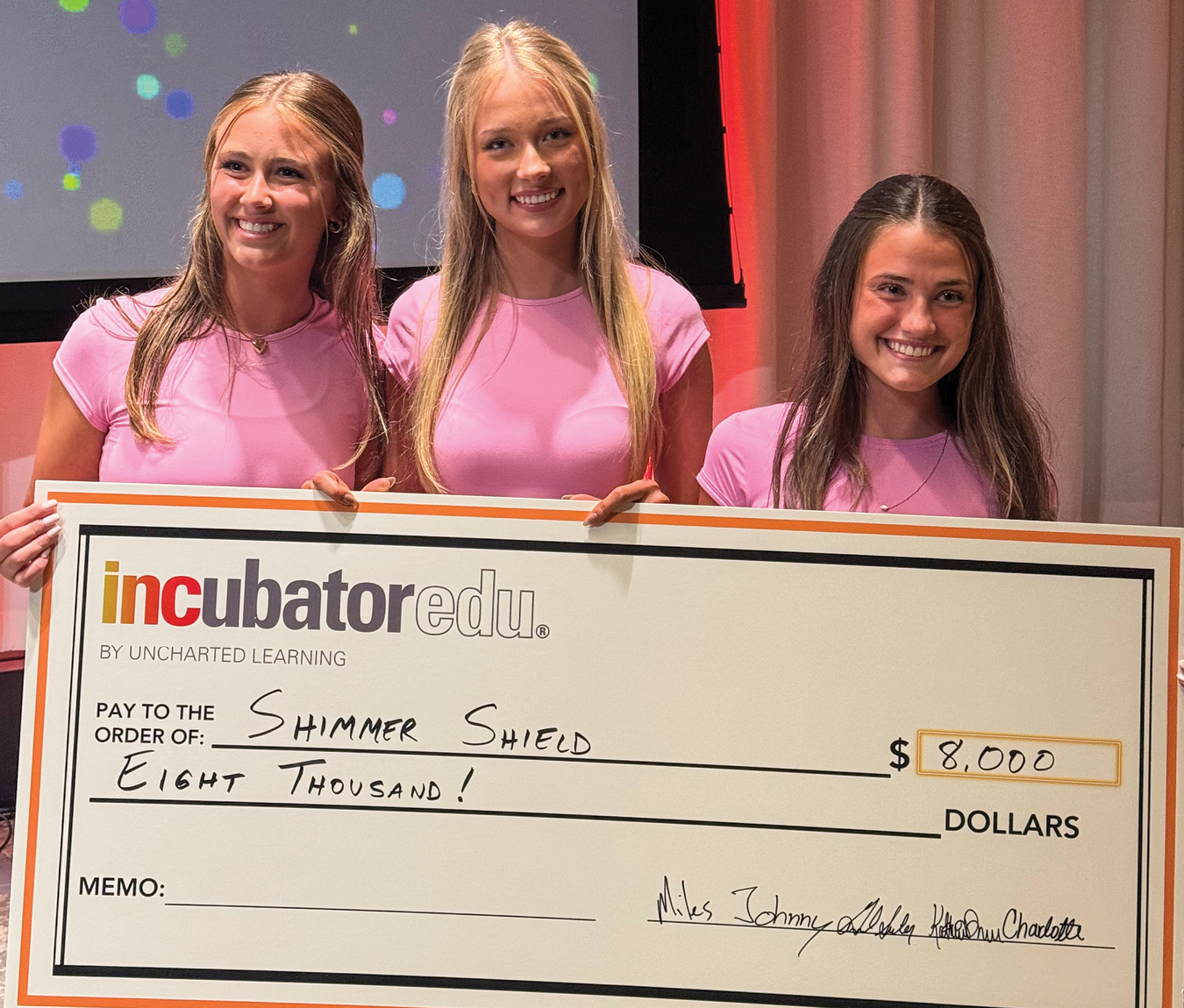 Three girls in pink shirts holding a large check