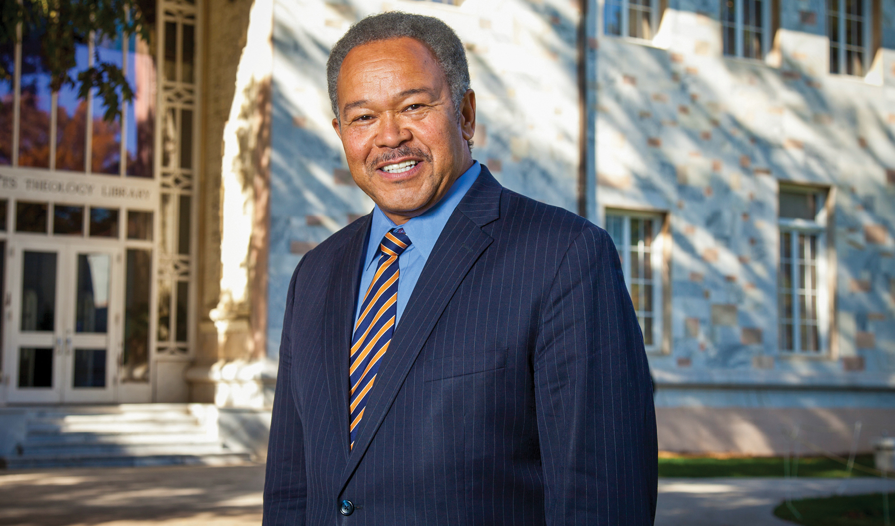 An outdoor headshot photo of a Black man in a suit and striped tie in front of a library