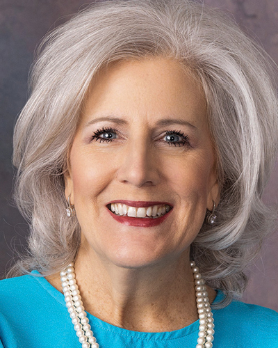 A white woman with gray hair smiling wearing a blue dress and pearls headshot