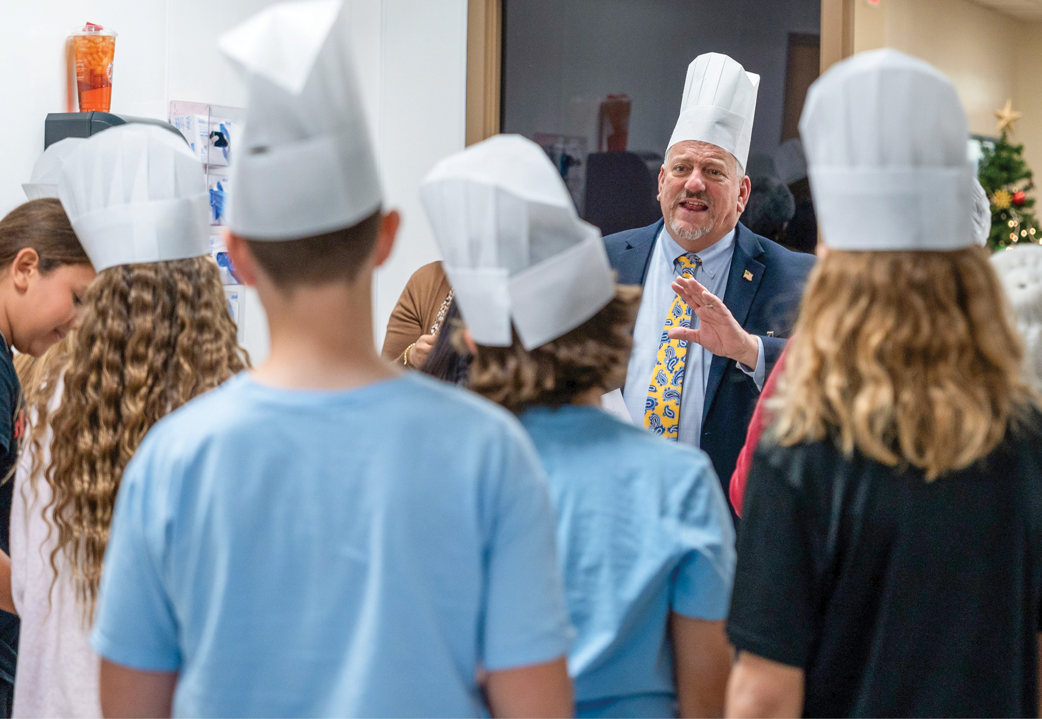 A man wearing a chefs hat talks to a group of students wearing chefs hats