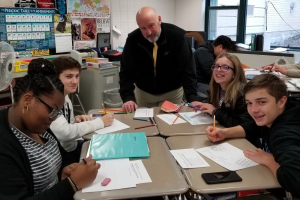 Todd Gazda stands at a cluster of desks with students in a classroom